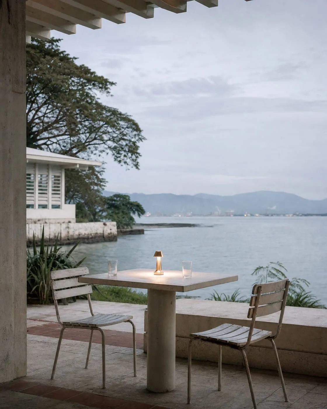 An outdoor table at dusk with the sky still pale and luminous, one small warm lamp just switched on, two empty glasses on the table, no people.