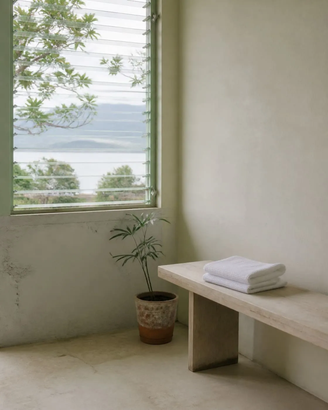 A pale room with an open louvred window, a folded white towel on a plain wooden bench and a small potted plant nearby, bright overcast daylight, no people.