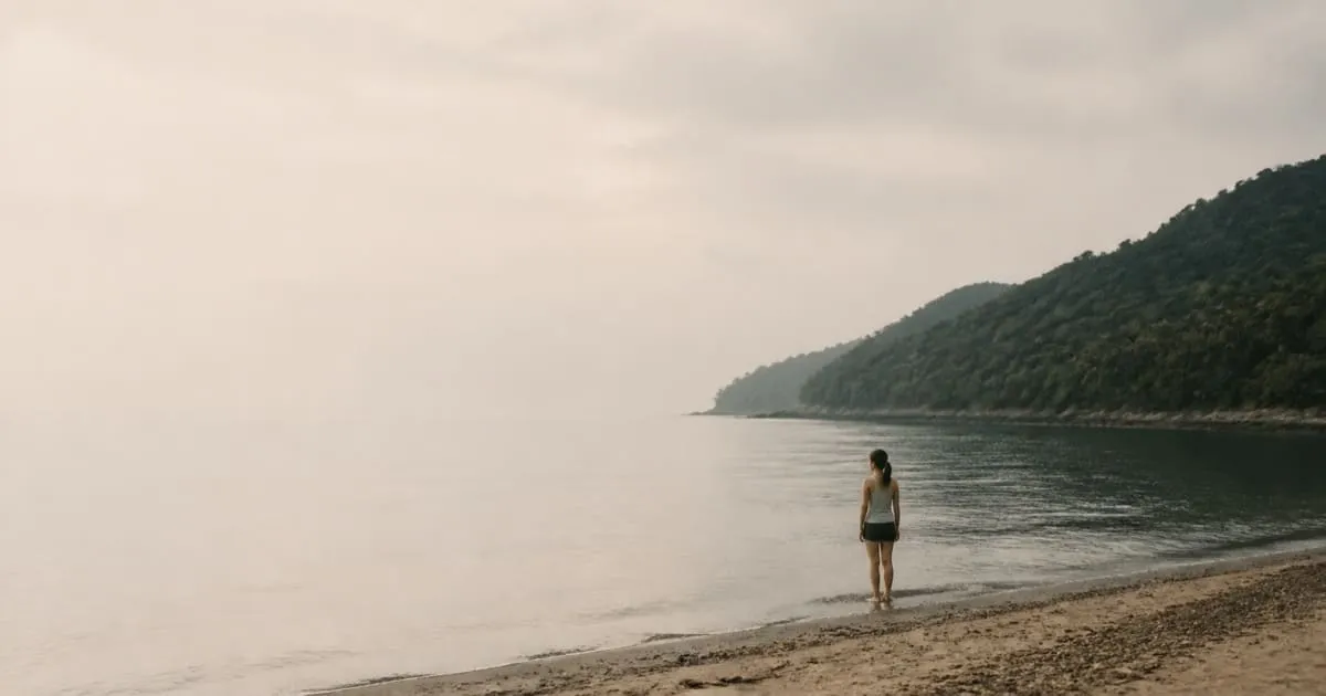 A person seen from behind at the edge of a quiet bay in early morning haze, forested headland to the right, wide open sky and sea filling the frame.