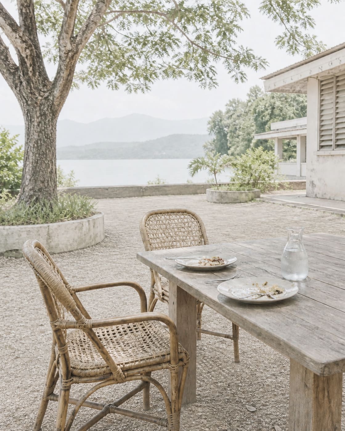 An outdoor wooden table under dappled acacia shade with two plates of food and a glass jug of water on it, two empty rattan chairs pulled out, no one seated, no hands visible.