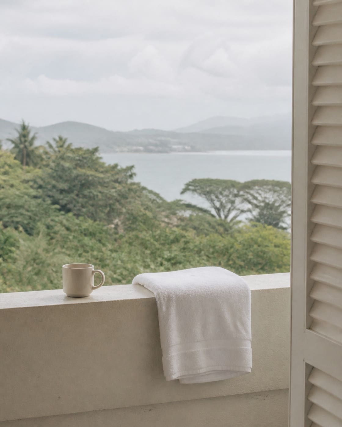 A folded white towel draped over a balcony railing beside a plain ceramic cup, pale louvred shutters to one side, a soft-focus view of forest and pale grey-green bay water beyond, overcast daylight, no people.