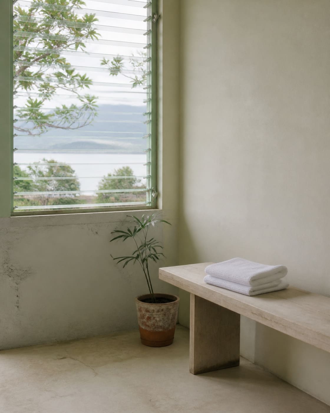 A pale room with an open louvred window, a folded white towel on a plain wooden bench and a small potted plant nearby, bright overcast daylight, no people.