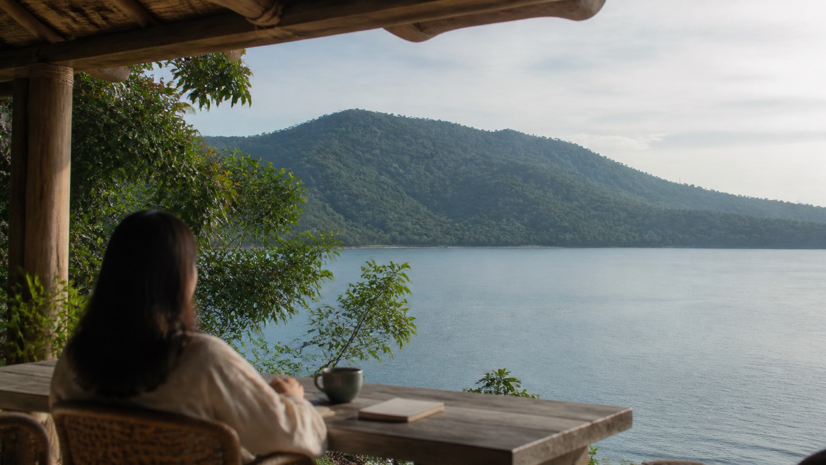 A person seen from behind sits with a warm drink on a shaded balcony overlooking the calm water and forested hills of Subic Bay.
